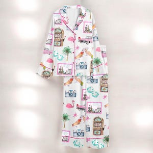 Puede incluir: Conjunto de pijama blanco con un estampado repetido de flamencos rosas, coches antiguos, m&aacute;quinas tragamonedas, c&aacute;maras, dados y palmeras. El conjunto incluye una camisa con botones con ribete rosa y pantalones a juego.