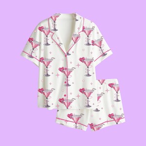 May include: A white short-sleeve button-down pajama shirt and matching shorts set featuring a repeating pattern of pink glittery cocktail glasses with heart-shaped garnishes and small four-pointed star accents, finished with pink piping trim.