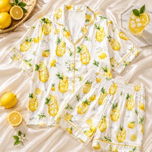 May include: A white short-sleeve button-down pajama shirt and matching elastic-waist shorts set featuring a repeating pattern of yellow lemon slices, green leaves, and glasses filled with iced lemonade and citrus garnishes with yellow piping.