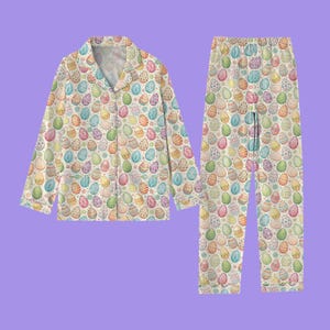 May include: A two-piece pajama set featuring a repeating pattern of colorful Easter eggs on a cream background. The long-sleeved top has a button-down front and a notched collar. The matching pants have an elastic waistband.