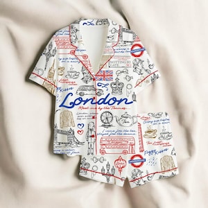 May include: A white London-themed pajama set featuring a short-sleeved shirt and shorts, both with red piping. The print includes illustrations of London landmarks such as the London Eye, a red double-decker bus, and the Union Jack flag.