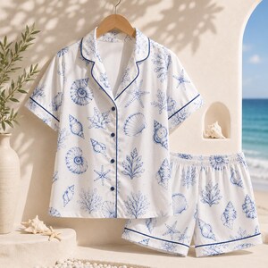 May include: A white pajama set featuring a blue seashell, starfish, and coral print. The short-sleeve button-down shirt has a notched collar with blue piping, and the matching shorts feature an elastic waistband with blue piping at the hem.