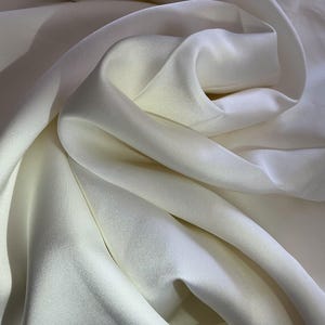 May include: A smooth, off-white fabric with a soft sheen and a dense, opaque weave. The material is draped in deep, flowing folds that create subtle highlights and shadows across the surface, emphasizing the fluid texture of the textile.