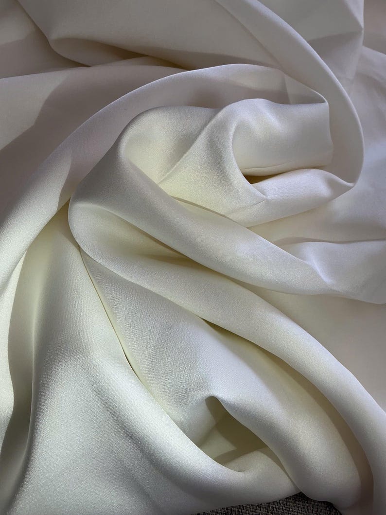 May include: A smooth, off-white fabric with a soft sheen and a dense, opaque weave. The material is draped in deep, flowing folds that create subtle highlights and shadows across the surface, emphasizing the fluid texture of the textile.