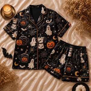 May include: A black short-sleeve button-down pajama shirt and matching shorts set featuring a repeating pattern of white ghosts, orange pumpkins, black cats, spiderwebs, stars, crescent moons, bats, and coffee pots with orange piping trim.