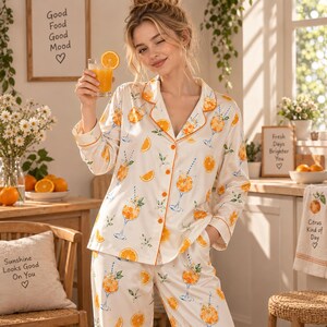 May include: A two-piece pajama set featuring a button-down long-sleeve shirt and matching pants. The fabric is cream-colored with an all-over print of orange slices, citrus cocktails in stemmed glasses, and green leaves, finished with orange piping.