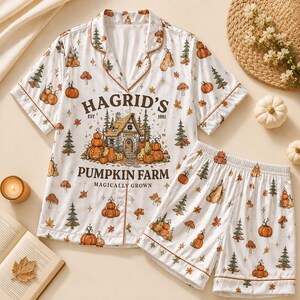 May include: A white short-sleeve button-down pajama shirt and matching shorts set featuring a repeating pattern of pumpkins, pine trees, and mushrooms. The shirt displays a central graphic of a cottage with the text HAGRID'S PUMPKIN FARM EST 1981.