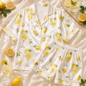 May include: A white satin pajama set featuring a short-sleeved button-down shirt and matching shorts. The fabric is decorated with a repeating pattern of yellow lemon slices, green leaves, and stemmed glasses filled with a yellow citrus beverage.