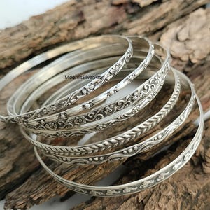 May include: A set of silver bangles with detailed floral and leaf designs. The bangles are displayed on a piece of wood, highlighting their intricate patterns. The jewellery is made of silver and features various embossed patterns.