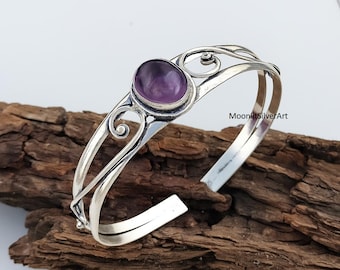 May include: A silver cuff bracelet with an oval, purple gemstone. The bracelet features a double band design with decorative swirls near the gemstone. The gemstone is set in a silver bezel. The bracelet is displayed on a piece of wood.