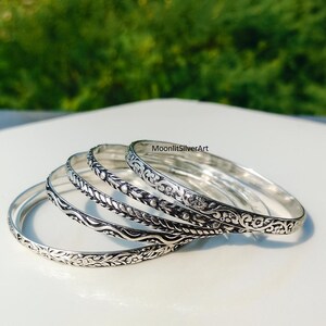 Op de afbeelding: Een verzameling zilveren armbanden met ingewikkelde bloemen- en bladerpatronen. De armbanden worden getoond op een wit oppervlak, met een vage groene achtergrond. De armbanden variëren in ontwerp en tonen gedetailleerd vakmanschap.
