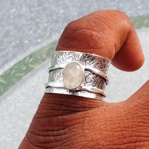 Puede incluir: Un anillo plateado con una piedra ovalada de color rosa claro en el centro. El anillo tiene un diseño floral y una banda giratoria. El anillo se lleva en un dedo.