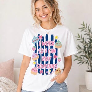 May include: A white short-sleeve t-shirt featuring a graphic print of dark blue vertical stripes, pink text that reads I WORK SO I CAN BUY SQUISHIES, and various colorful illustrations of small smiling pastries, strawberries, bows, and stars.