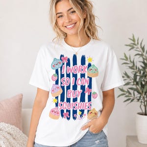 May include: A white short-sleeve t-shirt featuring a graphic print of vertical navy blue brush strokes, pink text that reads I WORK SO I CAN BUY SQUISHIES, and various colorful illustrations of small smiling squishy toys, bows, and stars.