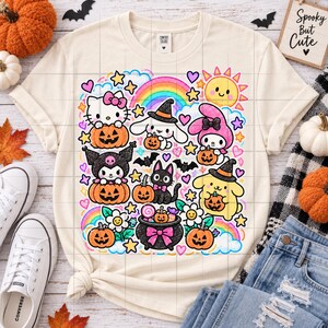 May include: A cream-coloured t-shirt featuring a colourful graphic print of various cartoon characters, including a white cat with a pink bow, a black cat, and a yellow dog, all holding orange jack-o'-lanterns, surrounded by rainbows, stars, and hearts.