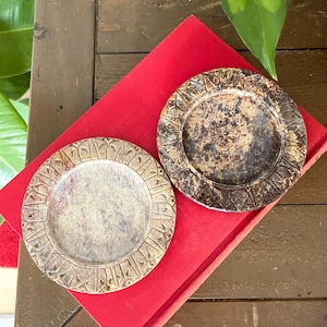 May include: Two circular stone coasters featuring a carved petal pattern along the outer rim. One coaster displays a light beige and grey mottled surface, while the other exhibits a darker, deep brown and black mottled finish with a textured appearance.