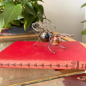 May include: A decorative insect figurine crafted from silver-toned wire, featuring a dark iridescent marble-like body and a smaller amber-colored glass sphere for the head, with thin wire antennae and legs, resting on a bright red hardcover book.