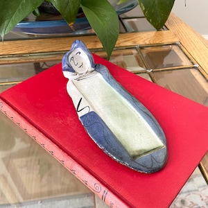 May include: A long, oval-shaped ceramic tray featuring a hand-painted figure at one end with a blue hood and a simple facial expression. The center of the tray is a smooth, light cream color, while the outer edges are finished in a deep blue glaze.