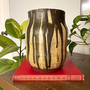 May include: A cylindrical ceramic vase with a speckled cream-colored base and dark brown vertical drip glaze patterns. The vase features a wide, open top and a solid dark brown band at the bottom, resting on a red book with decorative black spine text.