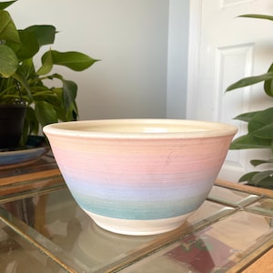 May include: A ceramic bowl featuring a horizontal gradient pattern with soft pastel bands of peach, pink, lavender, and light blue. The bowl has a wide, circular opening with a smooth, cream-colored interior and a tapered base.