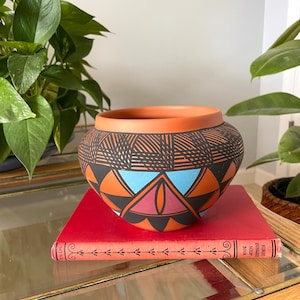 May include: A rounded terracotta pot featuring a matte black upper band with etched cross-hatch patterns and a lower section decorated with geometric triangles in shades of bright orange, light blue, and muted pink, resting on a red book.