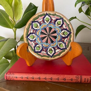 May include: A circular ceramic tile featuring a complex geometric star pattern in shades of pink, green, blue, and black with a gold-toned rim. The tile is displayed on a wooden stand, which is resting on a red book with decorative black line art.