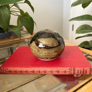 May include: A spherical ceramic object with a speckled tan and brown glaze featuring dark brown drip patterns, resting on a bright red book titled Elbert Hubbard Scrap Book with a decorative black line pattern along the spine.