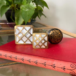 May include: Two square cabinet knobs featuring a brass lattice overlay with white mother-of-pearl inlays and one round black cabinet knob with a gold-toned filigree pattern, all resting on a red book with a black decorative spine design.
