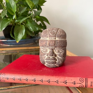 May include: A brown stone sculpture of a human head featuring stylized facial features and a decorative headband with geometric carvings. The sculpture has a textured, matte surface and is positioned on a red book with black decorative line patterns.
