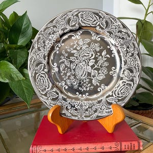 May include: A round, metallic silver-toned decorative plate featuring an intricate, embossed white floral pattern of roses and scrolling foliage across the rim and center. The plate is displayed upright on a wooden stand atop a red book.