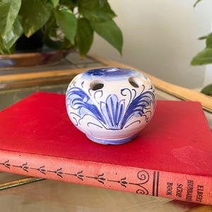 May include: A round, white ceramic flower frog featuring hand-painted blue floral and scroll motifs. The object has a flat top with multiple circular openings for stems and a slightly tapered base, resting on a red book titled Elbert Hubbard's Scrap Book.