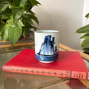 May include: A cylindrical ceramic tea cup featuring a white base with hand-painted blue ink illustrations of mountains and trees. The cup has a solid blue band around the base and a thin brown rim. It is resting on a red hardcover book.