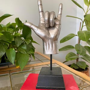 May include: A silver-colored hand sculpture displaying the American Sign Language sign for I love you. The hand is mounted on a thin black metal rod attached to a square black base, which is resting on top of a red book with visible text on the spine.