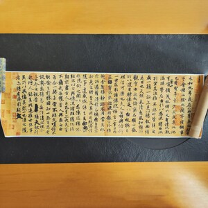 Pode incluir: Um pergaminho parcialmente desenrolado com caligrafia preta sobre um fundo amarelado. O pergaminho está sobre uma superfície preta. O texto está em uma língua asiática, com selos vermelhos.