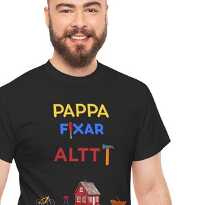Puede incluir: Camiseta negra con las palabras "PAPPA FIXAR ALTT" en amarillo, azul y rojo. Debajo del texto hay pequeñas ilustraciones de una bicicleta, una casa y un barco. La camiseta la lleva una persona.