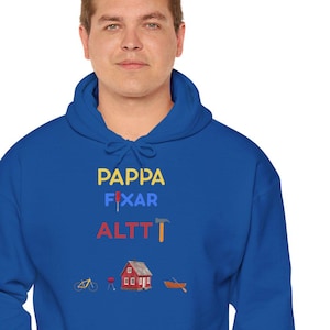 Puede incluir: Una sudadera con capucha azul real con el texto "PAPPA FIXAR ALTT" en amarillo, azul claro y rojo. Debajo, pequeñas ilustraciones de una bicicleta, una casa y un barco. La sudadera tiene un bolsillo delantero y una capucha con cordón.