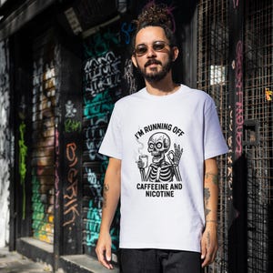 Puede incluir: Camiseta blanca con un diseño gráfico en blanco y negro de un esqueleto sosteniendo una taza de café y el texto "I'M RUNNING OFF CAFFEINE AND NICOTINE". La persona está de pie frente a una pared cubierta de grafitis.
