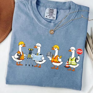 May include: Blue jean-colored t-shirt featuring a whimsical design of four geese dressed as construction workers. Each goose wears a hard hat and safety vest, holding tools like a clipboard, blueprints, and a stop sign. A gold necklace with a pendant is draped across the shirt.