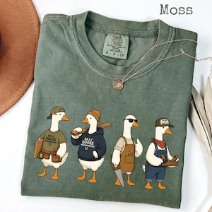 Peut inclure: Un t-shirt vert mousse avec un motif de quatre oies de dessin animé habillées en menuisiers. Les oies portent des chapeaux, des tabliers et des sweats à capuche avec le texte Silly Goose Woodworking. Le t-shirt a un col rond et une étiquette Comfort Colors.