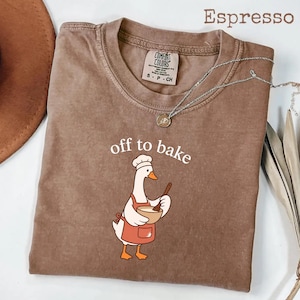 May include: An espresso-colored t-shirt featuring a graphic of a white goose wearing a chef hat and an orange apron, holding a wooden spoon and a mixing bowl. The text above the goose reads off to bake. The shirt has a Comfort Colors brand label.