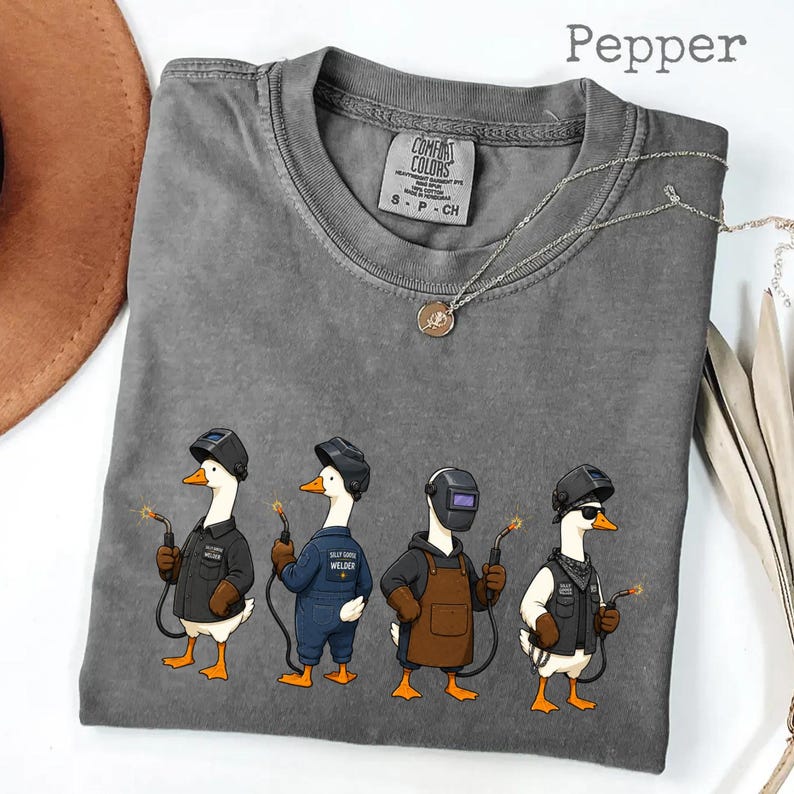 May include: A pepper-colored t-shirt featuring a graphic of four geese dressed as welders, each wearing a welding helmet and holding a welding torch. The shirt has a crew neckline and a Comfort Colors brand label printed on the inside collar.
