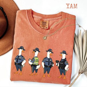 May include: A terracotta-colored short-sleeve t-shirt featuring a graphic of four geese dressed as state troopers. The geese wear blue uniforms, hats, and tactical gear. The text YAM is visible in the top right corner of the image.