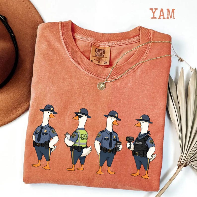 May include: A terracotta-colored short-sleeve t-shirt featuring a graphic of four geese dressed as state troopers. The geese wear blue uniforms, hats, and tactical gear. The text YAM is visible in the top right corner of the image.