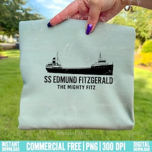 Peut inclure: Un t-shirt bleu clair avec une silhouette noire du SS Edmund Fitzgerald, un cargo des Grands Lacs. Le texte "SS EDMUND FITZGERALD" et "THE MIGHTY FITZ" sont imprimés sous l'image du navire. Le t-shirt est tenu par une main.