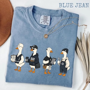 May include: A blue jean colored t-shirt featuring a graphic of four geese dressed as industrial engineers. The geese wear various work gear including hard hats, safety goggles, and overalls, while holding tools, clipboards, and tablets.