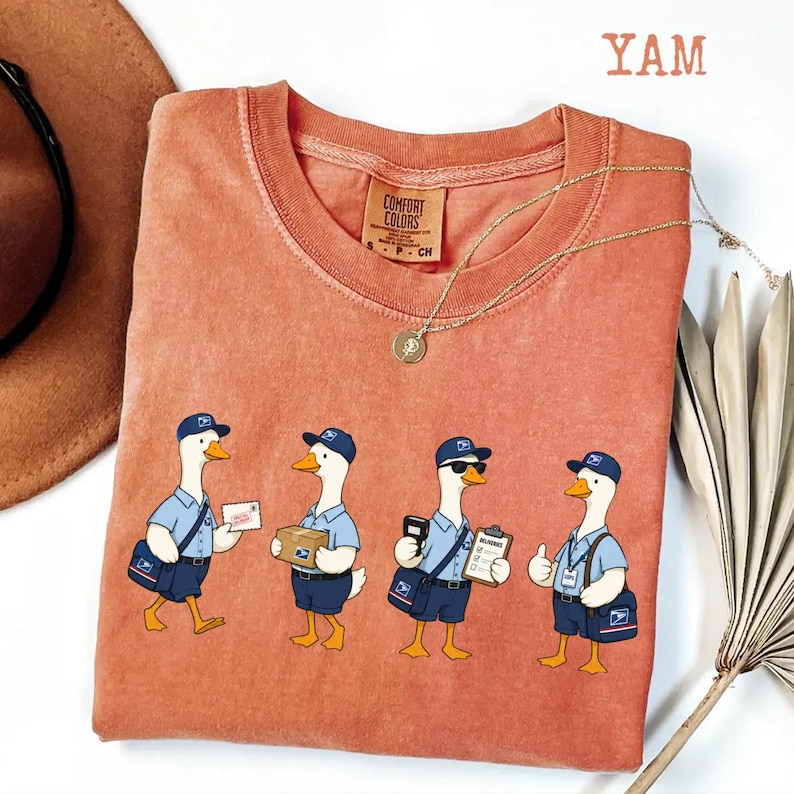 Puede incluir: Una camiseta Comfort Colors de color coral con un dise&ntilde;o caprichoso de cuatro gansos de dibujos animados vestidos como carteros. Cada ganso se representa en una pose diferente, llevando correo, paquetes y portapapeles. La palabra "YAM" est&aacute; impresa en la esquina superior derecha.