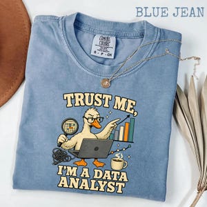 May include: A blue jean coloured t-shirt featuring a graphic of a duck wearing glasses, holding a magnifying glass, and working on a laptop. The text reads, "TRUST ME, I'M A DATA ANALYST," accompanied by a bar chart, a coffee mug, and tangled wires.