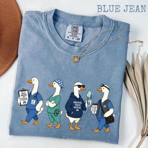 May include: Blue jean colored t-shirt with a graphic of four geese dressed as respiratory therapists. The geese are wearing scrubs, hoodies, and sunglasses, and carrying medical equipment. The text "Breathe, Treat, Repeat" is on one hoodie.