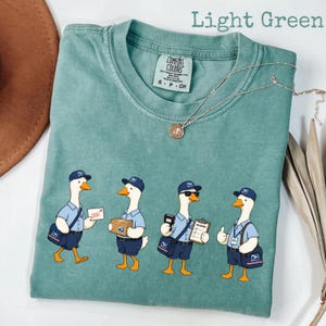 Puede incluir: Camiseta verde claro con un gr&aacute;fico de cuatro gansos de dibujos animados vestidos de carteros. Cada ganso lleva un uniforme azul, gorra y bandolera. El texto "Light Green" est&aacute; en la parte superior de la imagen.