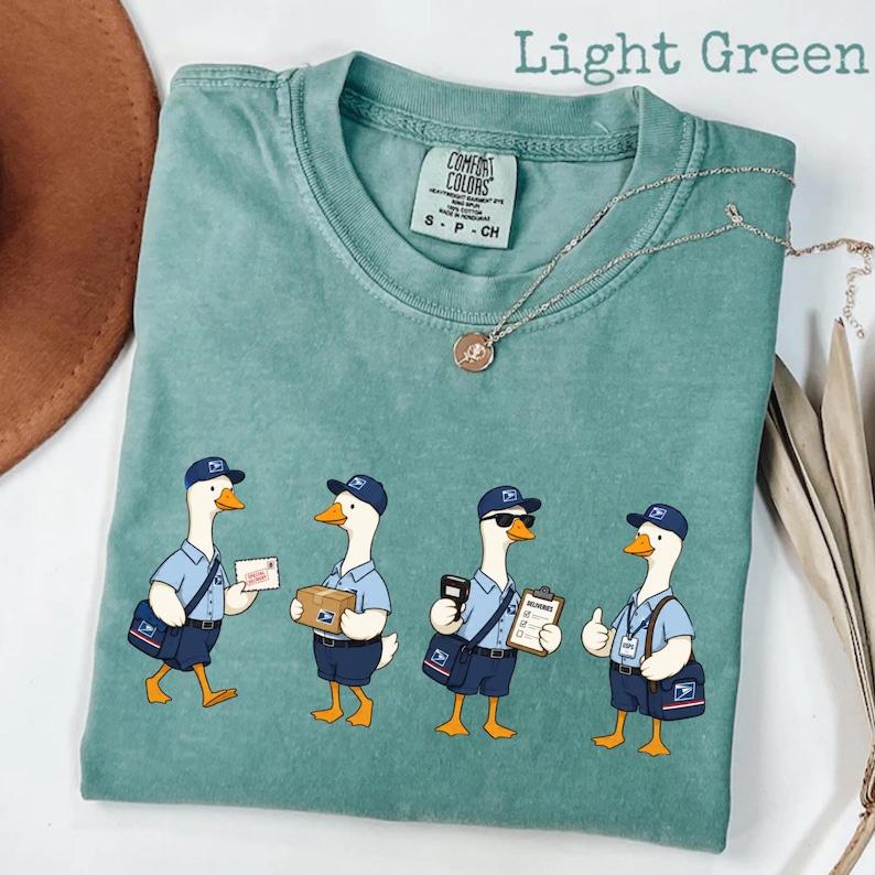 Puede incluir: Camiseta verde claro con un gr&aacute;fico de cuatro gansos de dibujos animados vestidos de carteros. Cada ganso lleva un uniforme azul, gorra y bandolera. El texto "Light Green" est&aacute; en la parte superior de la imagen.
